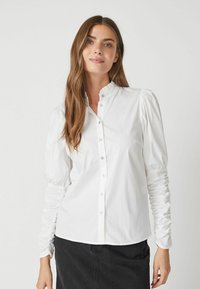 Camicia bianca con colletto arricciato e maniche lunghe e gather. Realizzata in un tessuto morbido, presenta una vestibilità a taglio sartoriale e una leggera lucentezza.