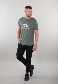 Zelené bavlněné tričko s bílým logem "Alpha Industries", spárované s černými cargo kalhotami s kapsami a texturovanými černými teniskami.