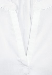 Blusa in tessuto bianco con scollatura a spiovente, con dettagli di cucitura ben visibili e una texture liscia. Adatta per essere indossata a strati o per un look informale.