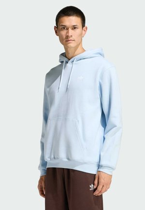 Mężczyzna w jasnobłękitnej bluzie Adidas z kapturem i ciemnobrązowych spodniach dresowych Adidas, stojący na tle jednolitego jasnego tła.