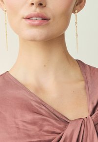 Gold chain earrings featuring circular links, hanging just below the earlobe, paired with a mauve, textured fabric top.