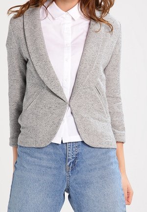 Femme portant un blazer gris clair texturé sur une chemise blanche boutonnée et un jean bleu, debout devant un fond uni.