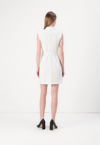White sleeveless dress with a belted waist, round collar, and smooth texture; paired with black high-heeled shoes. Rear view.