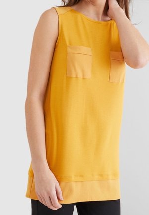 Haut jaune sans manches avec deux poches poitrine porté par une personne avec une main touchant son cou et l'autre sur le côté.