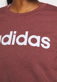 Personne portant un t-shirt bordeaux avec un texte blanc en gras montrant partiellement le nom de la marque "adidas" sur la poitrine.