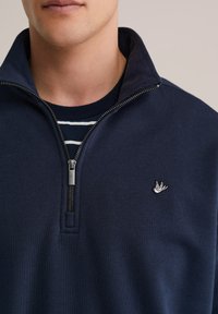 Tengerészkék pulóver magas gallérral, cipzárral és a mellkason hímzett madár logóval. Puha, texturált anyagból készült.