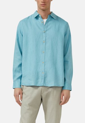 Mann, der ein hellblaues langärmeliges Hemd mit Knöpfen über einem weißen Unterhemd und hellbeigen Hosen trägt, steht vor einem einfarbigen Hintergrund.