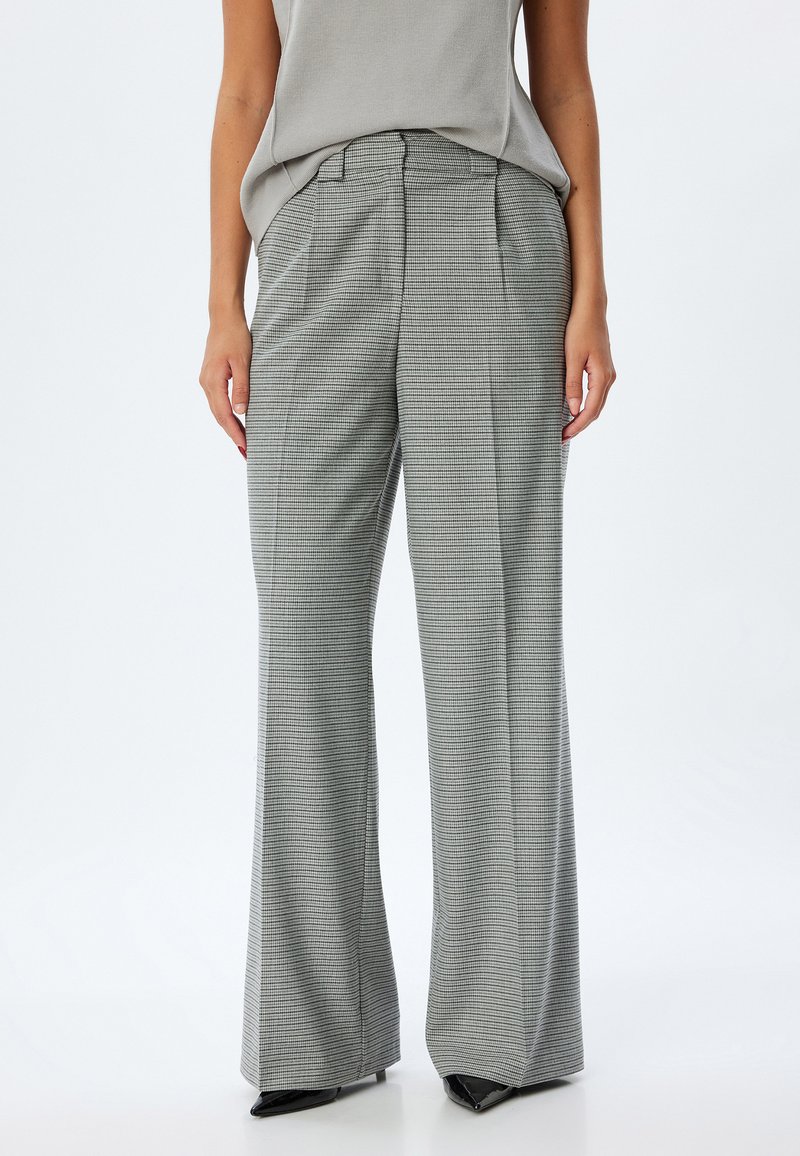 Pantalons larges à motif à carreaux gris fabriqués en tissu texturé, avec une taille haute et des plis sur mesure. Accentués avec des chaussures noires.