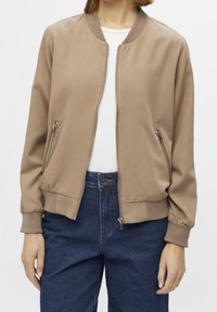 Braune Bomberjacke mit glatter Textur, mit Reißverschlusstaschen und gerippten Bündchen. Trägt man über einem weißen Hemd, kombiniert mit dunkelblauen Jeans.