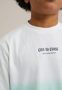 Balta medvilninė marškinėlių tunika su žaliu gradientiniu apvadu. Priekyje įspaustas jūrinio stiliaus logotipas "CITY TO COAST BLUE RIDGE DENIM".