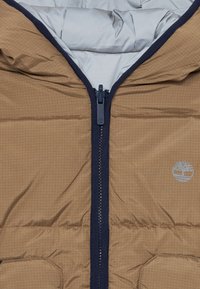 Doudoune marron avec une texture en grille, accents navy, fermeture éclair à l'avant et doublure grise. Comprend un logo et de grandes poches avant.
