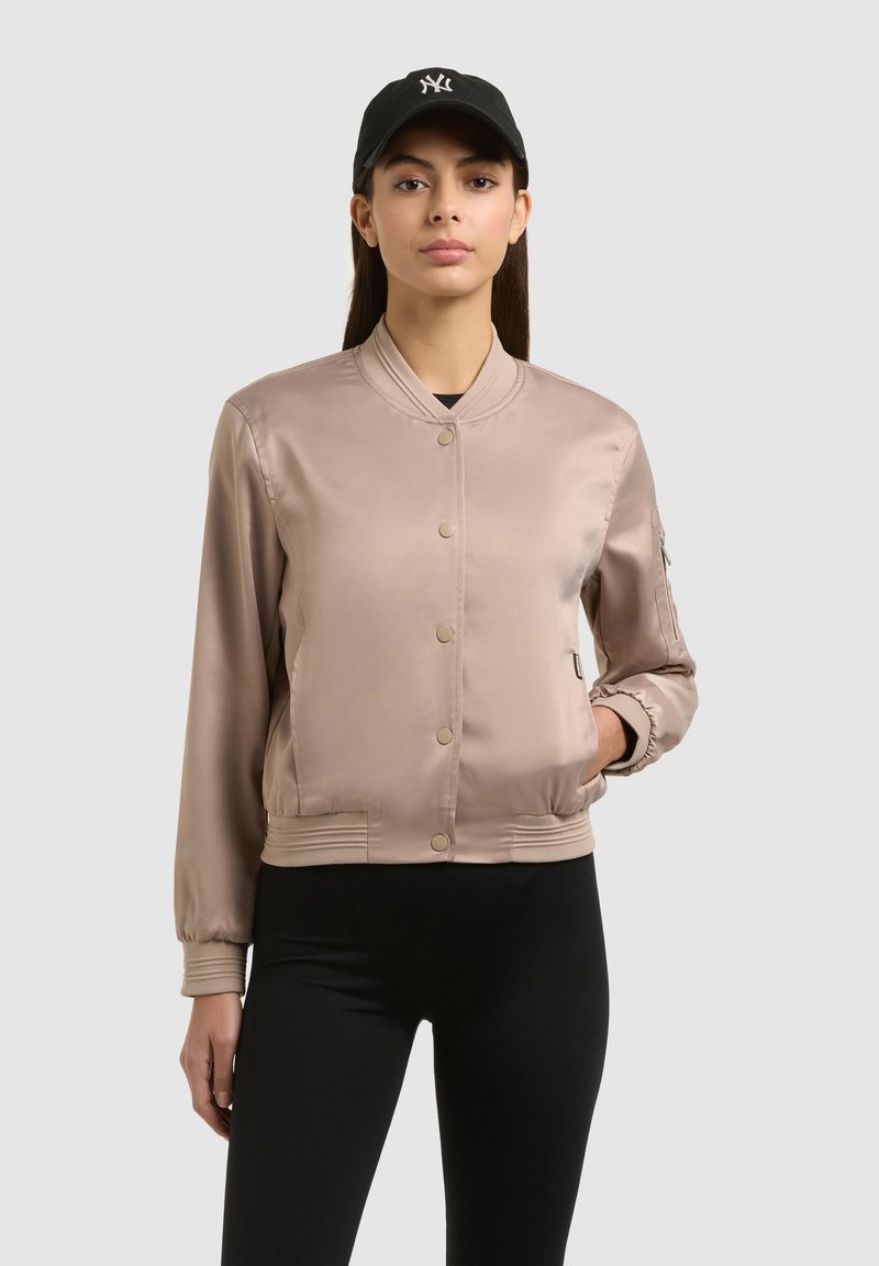 Femme portant un bomber en satin beige, des leggings noirs et une casquette noire, debout devant un fond uni gris clair.