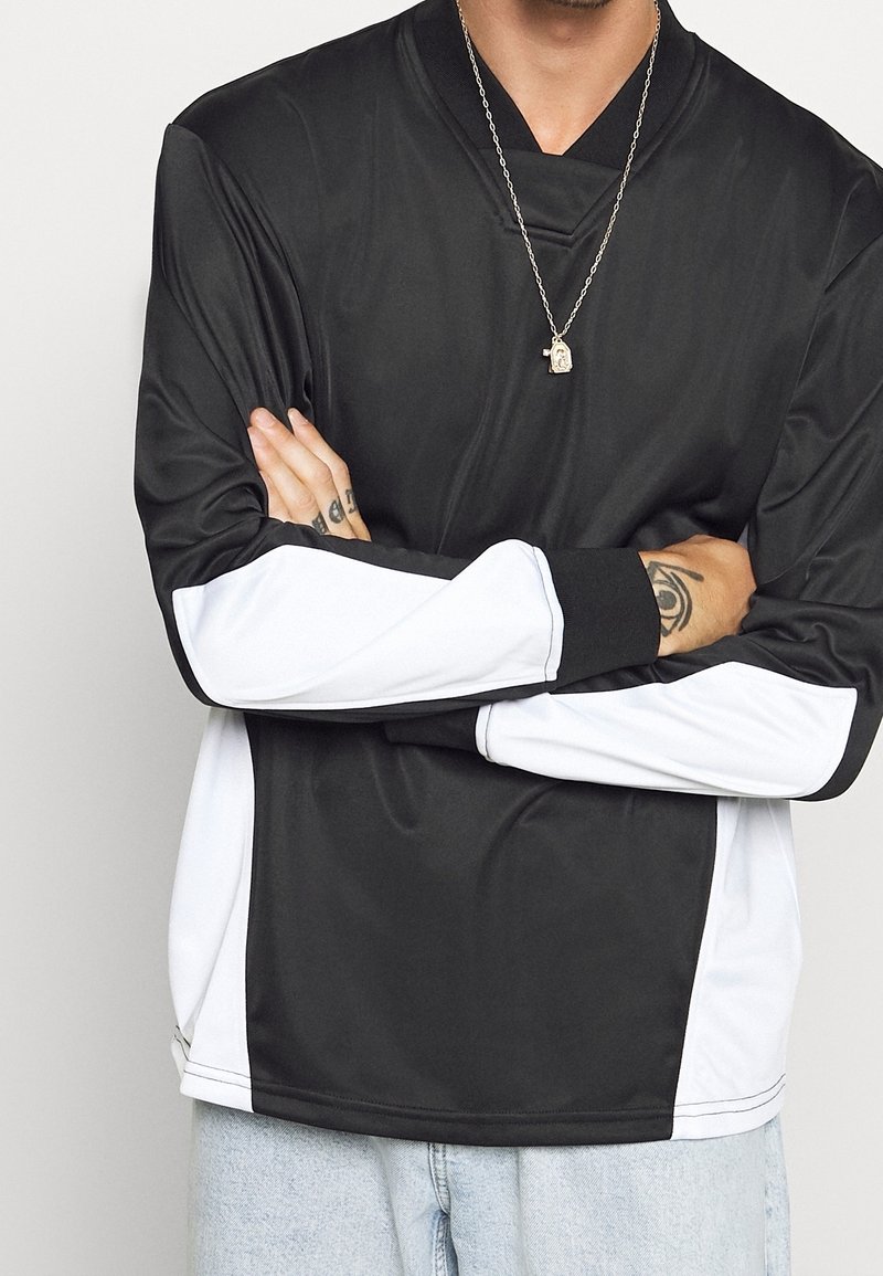 Haut à manches longues noir et blanc avec une texture lisse, doté d'une fermeture éclair au col et de panneaux latéraux blancs contrastants. Porté avec un jean clair.