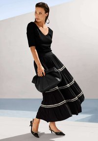 Schwarzes, figurbetontes Top kombiniert mit einem schwarzen, plissierten Maxirock mit weißen, gerafften Streifen. Die Accessoires umfassen eine schwarze Lederhandtasche und spitze Absatzschuhe.