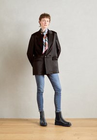 Crna blazer jakna s satenskim završetkom, svijetloplava košulja, šarena prugasta marama, plave traperice i teksturirane crne čizme do gležnja.