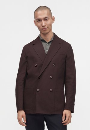 Mand iført en mørkebrun dobbeltradet blazer over en grå skjorte med krave mod en ensfarvet hvid baggrund.