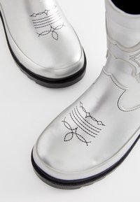 Sølvfarvede vandtætte støvler med sorte dekorative sømmønstre. Designet har en glat metallisk overflade og en sort sål.