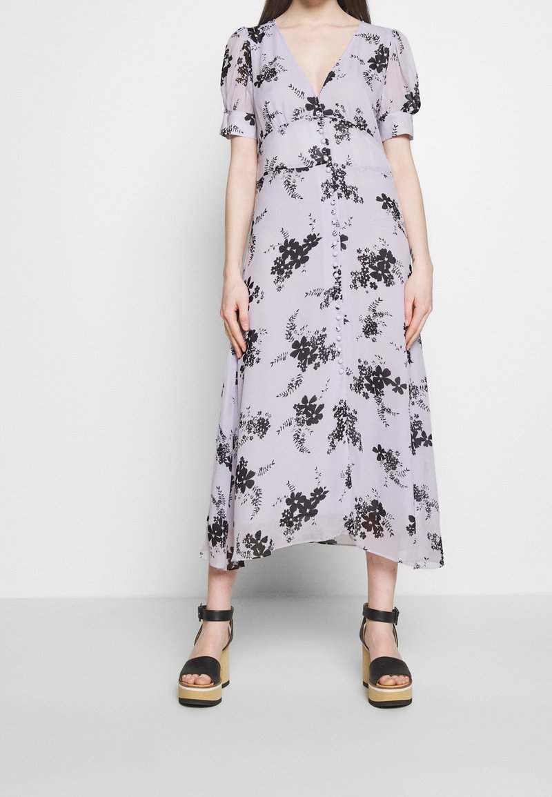 Femme portant une robe midi fleurie lavande avec manches bouffantes, détail boutonnière sur le devant et sandales compensées noires avec brides à la cheville.