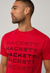 Camiseta de algodón roja con un diseño de texto negro repetido que dice "HACKETT". Cuello redondo, mangas cortas, textura suave y forma ajustada.