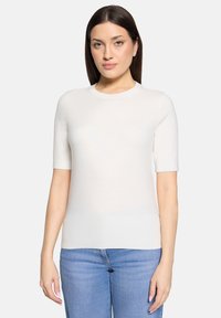 Pull léger à manches courtes de couleur crème avec un col rond et une texture lisse, associé à un jean bleu. Aucun motif ni accessoire visible.