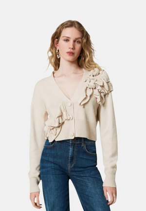 Donna che indossa un cardigan color crema con grandi volant in tessuto e jeans blu scuro, in piedi davanti a uno sfondo semplice.