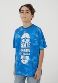 T-shirt blu tie-dye in cotone, con grafiche bianche di skateboard e la scritta "SKATE BOARDING LOS ANGELES." Maniche corte, vestibilità rilassata.