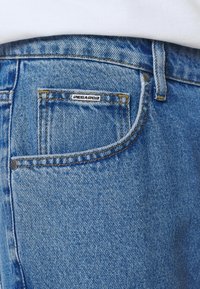 Jeans de mezclilla azul desvanecidos con un bolsillo delantero visible, costuras amarillas y una etiqueta plateada con la marca "PEGADOR" cerca de la parte superior.