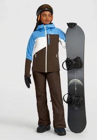 Bruine en blauwe snowboardjas met witte accenten, gecombineerd met bruine sneeuwbroeken. Zwarte snowboard met bindingen, gedragen met zwarte handschoenen en een helm.