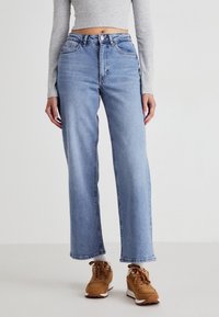 Femme portant un jean bleu clair droit, un top court gris à manches longues, des chaussettes blanches et des baskets marron, debout devant un fond uni.
