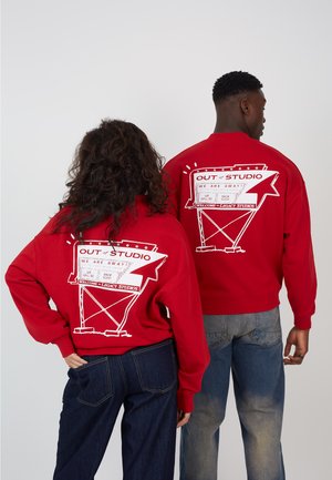 Due persone che indossano felpe rosse con una grafica bianca sul retro con la scritta "OUT of STUDIO WE ARE AWAY", in piedi fianco a fianco.
