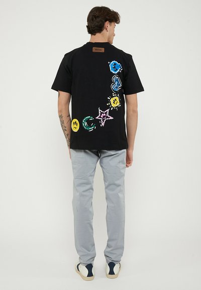 Man wearing black T-shirt with colorful smiley, star, and shape designs on back, gray pants, and white sneakers, standing facing away.