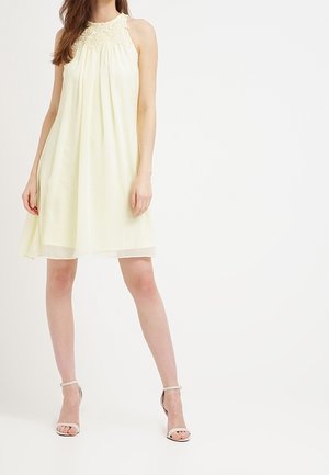 Femme portant une robe crème sans manches, longueur genou, avec un détail en dentelle au niveau du décolleté, associée à des talons blancs à bride cheville.