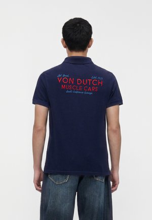 Mann trägt marineblaues Polo mit rotem und blauem Schriftzug „Von Dutch Muscle Cars South California Garage“ auf dem Rücken, kombiniert mit blauen Jeans.