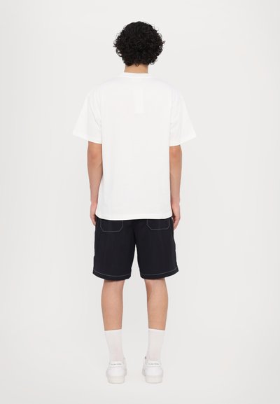 White cotton t-shirt with short sleeves, paired with black shorts featuring white stitching and a casual fit. Model stands facing away.