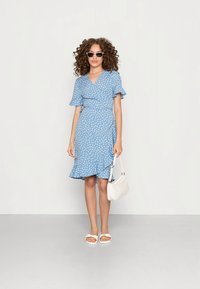 Svetlomodré zavinovacie šaty s bielymi bodkami, krátkymi nariasenými rukávmi a nariaseným lemom. Nosia sa s bielymi sandálmi a malou bielou kabelkou.