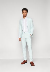 Traje azul claro con un diseño entallado, tejido texturizado, pantalones a juego y una camisa blanca de vestir. Zapatos de cuero marrón completan el atuendo.