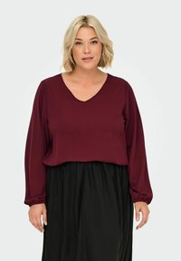 Burgundy long-sleeve top with a V-neck design and gathered cuffs, paired with a flowing black skirt. Soft fabric with a smooth texture.