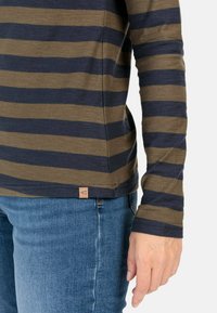 Langarmshirt mit horizontalen olivgrünen und navyblauen Streifen, aus weichem Stoff, seitlichem Etikettendetail, kombiniert mit blauen Jeans.