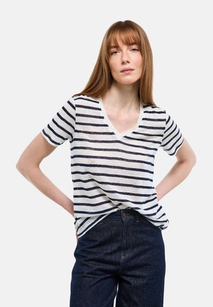 Jeune femme aux cheveux rouges lisses portant un t-shirt à col en V blanc rayé de bleu marine et un jean bleu foncé, debout les mains sur les hanches.