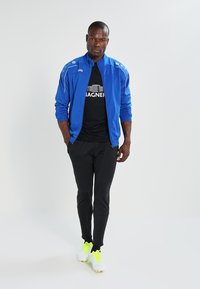 Blauwe sportjack met ritssluiting, zwart T-shirt met logo, en zwarte slim-fit broek. Witte en neongele sportschoenen.