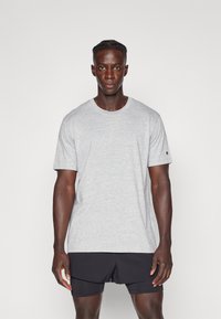 Graues Kurzarm-T-Shirt aus Baumwollstoff, mit rundem Halsausschnitt und einem Logo auf dem linken Ärmel, kombiniert mit schwarzen Shorts.