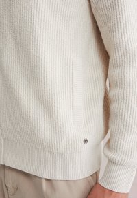 Kreemjas soonikkoega kampsun, millel on tekstuurse pinnaga kangaroo-tasku ja väike logo detail ääre lähedal.