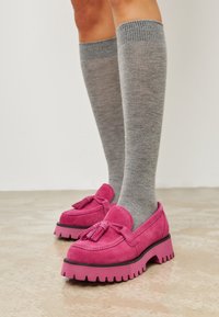 Rosa Wildleder Slipper mit Quastenverzierung und dicken, gezahnten Sohlen, kombiniert mit grauen knielangen Socken aus einem strukturierten Material.
