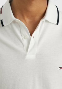 Bílá polo košile s tří knoflíkovým zapínáním a kontrastním límcem se červenými a černými pruhy. Emblém na prsou. Jemná textura.