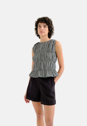 Joven con el pelo corto y rizado, lleva una camiseta sin mangas de cuadros en blanco y negro y pantalones cortos negros, de pie con las manos en los bolsillos.