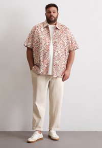 Chemise à manches courtes à motif floral crème et rouge, assortie à un t-shirt blanc, un pantalon beige clair et des baskets blanches à semelles en caoutchouc.