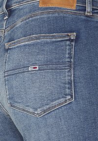Blå denimjeans med en rektangulär bakficka, med röd och vit logobroderi. Läderlapp ovanför fickan. Slät textur.