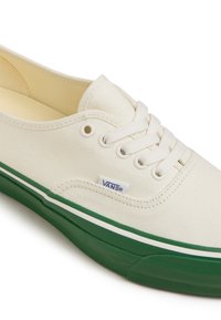 Sneaker bianca in tela con suola in gomma verde e lacci bianchi. Presenta un'etichetta con logo blu e cuciture distintive lungo i bordi.