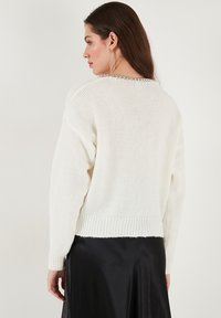 Valge koeheegeldatud särk ribatud äärise ja tekstuuriga mustriga. Kaelas on kettdetail. Tagasiside näitab pikki varrukaid.