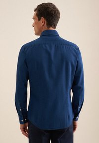 Uomo che indossa una camicia blu scuro con maniche lunghe e bottoni, in piedi rivolto di spalle su uno sfondo semplice.
